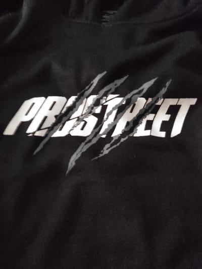 The image displays a close-up view of a black fabric garment, identified as a hooded sweatshirt, observed in Linkebeek, Belgium. The central front area of the garment features a prominent horizontally oriented graphic. This graphic consists of the word "PROSTREET" rendered in solid white, sans-serif, uppercase letters. Five distinct, diagonal claw-mark graphics, colored in a light grey with darker grey outlines and internal striations, are superimposed over the white text, appearing to tear through portions of the letters. The fabric exhibits minor creasing, and the lighting is diffuse, casting no harsh shadows. No individuals, actions, or interactions are present within the frame.