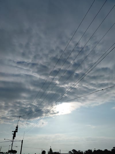 The image depicts a cloudy sky with power lines in the foreground. The sun is partially visible, suggesting it is daytime. The scene is likely outdoors, possibly in a semi-urban or rural area given the presence of power lines and distant silhouettes of trees. There is no discernible activity or event taking place; the focus is on the natural elements and infrastructure. The clouds appear dense and varied, with some areas of brightness where the sun's rays are breaking through. The overall mood is somewhat dramatic due to the interplay of light and shadow on the clouds. No text is visible in the image.