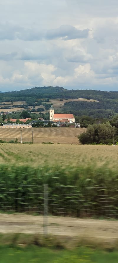 In the rural landscape of Loosdorf, Austria, a prominent church with a tall, green-domed spire stands out against a backdrop of rolling, tree-covered hills. The church features a long, red-tiled roof and pale yellow walls. In front of the church, a cluster of smaller buildings forms a village, with some modern, elongated structures visible to the left, possibly a school or administrative complex. The foreground of the image is dominated by agricultural fields. A row of tall green plants, likely corn, is blurred due to motion, suggesting the photo was taken from a moving vehicle. Behind this, a dry, harvested field is visible, with distinct rows indicating past cultivation. The sky above is filled with dramatic, white and grey clouds. The scene is peaceful and pastoral, characteristic of the Austrian countryside.