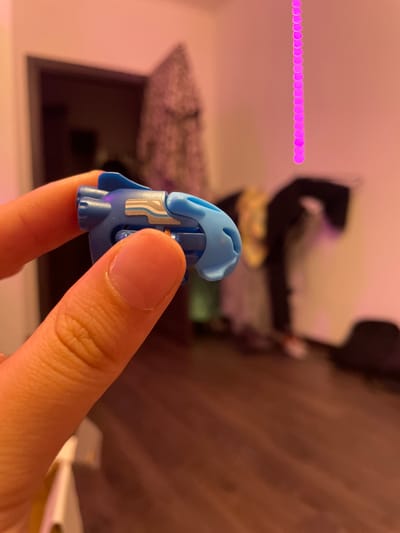 The image shows a close-up of a person's finger holding a blue plastic toy or component. The background is out of focus, showing a room with a dark doorway, a patterned fabric hanging on the wall, and a blurred figure in the distance. A string of pink, circular lights is visible on the right side of the image. The lighting suggests an indoor environment with artificial illumination, casting a warm, pinkish hue. There is no indication of the location being in Borisova Griva Massiv, Russia.