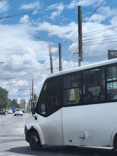 The image shows a white bus on a city street, seen from the right side. The bus is partially obscuring the view of the road ahead, where several other vehicles, including cars and a truck, are visible. In the background, there are apartment buildings and trees, along with utility poles and overhead wires against a blue sky with white clouds. The scene appears to be captured during daytime. The presence of utility poles and the general urban architecture suggest this is likely a residential or commercial area within a city. There is no specific information in the image to confirm the location as Malaya Valeyevka, Russia.