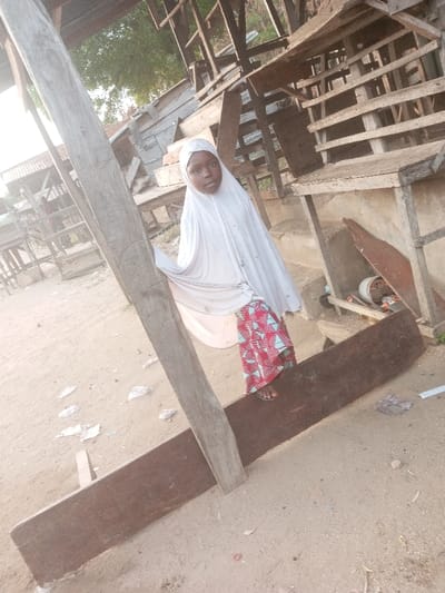 A young person wearing a white hijab and a patterned skirt stands near weathered wooden structures. One foot rests on a dark, elongated wooden plank. The scene is outdoors, with sandy ground and hints of foliage in the background. The wooden construction is exposed and seemingly unfinished, adding a rustic and somewhat derelict feel to the setting. The overall impression is one of a modest environment.