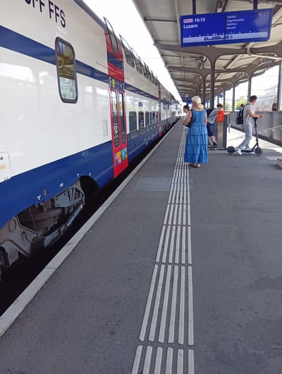 A train is stopped at a station platform. The train is white with blue and red accents and the letters "FFS" are visible on its side. A departure board above the platform indicates that train RE24, departing at 16:13 for Luzern, will stop in Reiden, Dagmersellen, Nebikon, and Wauwil, with track assignments A through H. Several people are visible on the platform. In the foreground, a woman in a long blue dress stands facing away from the camera. Behind her, a young person is on a scooter. Another person with a backpack is further down the platform. The scene appears to be a sunny day. The textured ground at the edge of the platform suggests it is designed for visually impaired individuals.