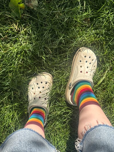 This is a top-down, close-up, high-angle shot of a person's legs and feet wearing beige Crocs and rainbow-striped socks, standing on green grass. The person is also wearing distressed denim shorts. The environment is outdoors with green grass and some fallen leaves, suggesting a park or a yard. The sun is shining, casting shadows. There is no indication of the location being in Kashka-Suu, Kyrgyzstan.