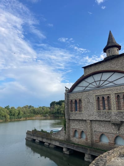 The image shows a stone building with a conical tower next to a body of water. The building has arched windows and a decorative stone wolf figure on its roof. The water is calm and reflects the building and the surrounding trees. The sky is blue with scattered clouds. There is a small airplane visible in the sky. The location is identified as Colibași, Moldova.