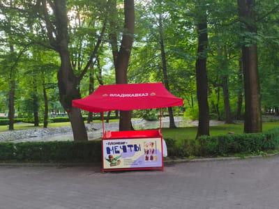 In Verkhnii Lars, Russia, a red awning stands over a small ice cream cart in a park. The awning displays Cyrillic text. The cart itself is decorated with images of ice cream and a cartoon character. Behind the cart, a neatly trimmed hedge borders a grassy area with trees. A stream or pond with rocky edges is visible further back, surrounded by more trees and greenery, suggesting a park or natural landscape. The ground in front of the cart is paved with grey interlocking stones. The overall scene depicts a calm outdoor setting on a day with diffused sunlight filtering through the leaves.