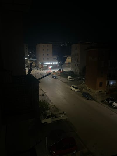 The image captures a street scene at night in Benevento, Italy. The perspective is from a height, looking down onto a street lined with multi-story buildings. Several cars are parked or driving on the street, including a white van, a white hatchback, a blue hatchback, a dark sedan, a dark SUV, and a white sedan. A small white flatbed truck is parked on the side of the road. The buildings show illuminated windows, suggesting occupation. Streetlights illuminate parts of the scene, casting shadows. There is some construction visible with orange fencing. A dark balcony railing and some architectural elements of a building are in the foreground on the left.