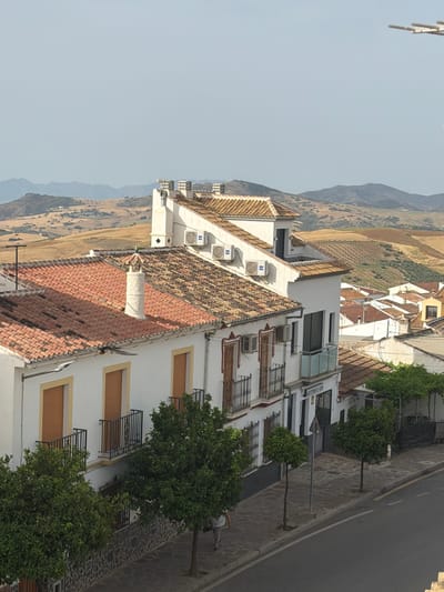 A high-angle, eye-level shot depicts a street scene in Villanueva De La Concepción, Spain, under a clear, hazy sky. The foreground is dominated by the upper floors of whitewashed buildings with terracotta tiled roofs. Several buildings feature multiple air conditioning units mounted on their exteriors, and ornate balconies with black wrought-iron railings. One building displays a sign reading "GESTORIA GNS." Lined trees, some appearing to be citrus trees with lush green foliage, grow along the sidewalk. A lone person, viewed from the back, walks along the sidewalk. The street is paved with cobblestones and asphalt. In the mid-ground and background, the landscape transitions to rolling hills covered in dry brush and olive groves, with distant mountains forming the horizon. An antenna array is visible in the upper right corner of the frame.