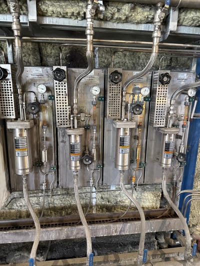 The image shows a section of industrial equipment, likely part of a water treatment or processing system, located in Krzepielów, Poland. The system consists of multiple vertical units, each containing a "Sample Cooler" manufactured by Sentry. These units are constructed of stainless steel and are mounted on a wall covered with insulation material. Various pipes, valves, pressure gauges, and flow meters are visible, connecting the different components. The overall environment appears to be an indoor industrial setting, with visible grates and some debris on the floor. There are no people present in the image.