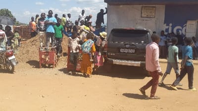 A crowd of people gathers in a street scene with a brown SUV parked. Some people stand on top of a dirt pile, others mill around the vehicle. A building with a sign can be seen behind the SUV. A motorcycle is parked on the left side of the image. Three people are walking in the foreground. The ground appears to be dirt or sand.
