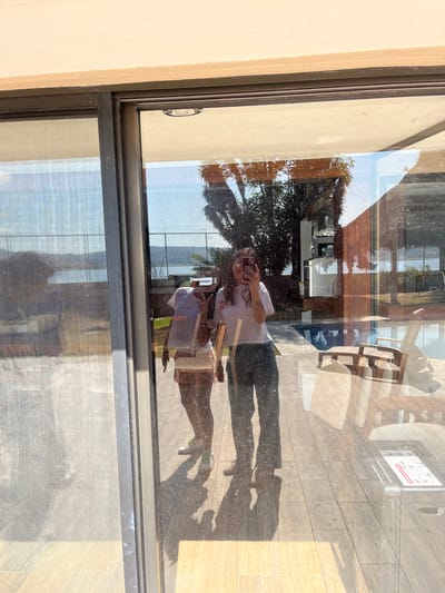 The image shows a reflection in a window in El Oasis Valsequillo, Mexico. The reflection includes a person holding a mobile phone, palm trees, a building, and outdoor furniture. The foreground shows the window frame and partially obscured view of the outdoors through the window. The person appears to be taking a picture of themselves in the reflection.