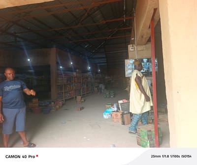 The image shows the interior of a general store in Langtang, Nigeria. The store has a corrugated metal roof supported by beams, with visible wiring. Fluorescent lights are attached to the walls, illuminating the shelves stocked with various items. A dark-skinned man in a blue t-shirt and shorts stands on the left. On the right, another man wearing a light-colored traditional outfit with a striped hat walks past a doorway. Behind him, there is a security monitor displaying multiple camera feeds. Boxes and various items are scattered on the dirt floor. There are shelves stocked with goods in the background. The boxes nearest the camera on the right include labels like "GONGONI CO. LTD" and "Mamuda".