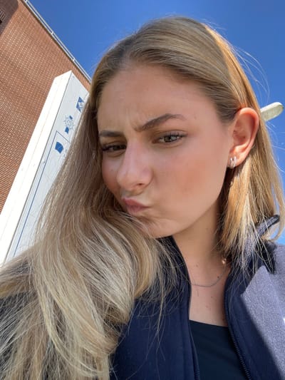 The image is a close-up, eye-level selfie of a young female with long, wavy blonde hair. She is directly facing the camera, making a "duck face" expression with puckered lips and a slight furrow in her brow. Her brown eyes are open, and she wears minimal visible makeup. She is dressed in a dark blue zippered fleece jacket over a black top, and a thin silver chain necklace is visible around her neck. Her right ear features two small hoop earrings; one appears to be plain silver, and the other is adorned with small, reflective stones.
The background indicates an outdoor, daylight setting. To the left of the subject, a section of a reddish-brown brick building is visible. Adjacent to the building, a tall, white vertical sign or pillar stands prominently. This sign features multiple blue stylized graphics and logos. One identifiable logo contains the letters "ESIC" and a distinct corporate emblem, while other blue graphics resemble academic crests or symbols. This signage, combined with the building architecture, identifies the location as the ESIC Business & Marketing School campus in Pozuelo de Alarcón, Spain. Above and to the right of the subject, a clear blue sky is present, with a small, white, indistinct object partially visible against it.