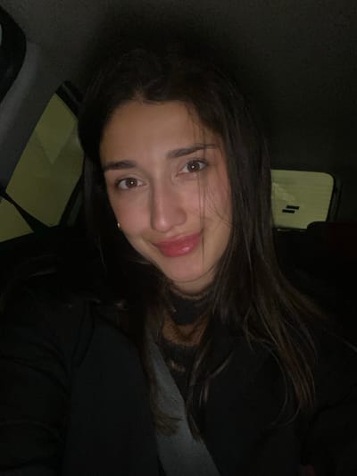 The scene depicts a female individual inside a vehicle, illuminated by an immediate light source, likely a camera flash or phone screen, against a dark, low-light background. The precise location within the city of Đurmani, Montenegro, cannot be determined from the visual information provided.
The primary subject is a young adult female, positioned centrally, facing directly towards the camera. She possesses long, dark brown hair, parted on the left, with some strands falling over her forehead and shoulders. Her complexion is olive, and she has brown eyes. A faint gold hoop earring is visible on her left earlobe. Her lips are coated with a glossy, light pink substance, and she displays a slight, closed-mouth smile. She is wearing a dark-colored, possibly black, outer garment over a dark, textured or patterned high-neck top. A grey seatbelt diagonally traverses her torso from her left shoulder towards her right hip.
The immediate environment appears to be the interior of an automobile. Blurred light sources, predominantly yellow-green, are visible through the vehicle's windows on the left side, suggesting an outdoor setting at night or in a poorly lit area. The rear window is visible behind her right shoulder, showing indistinct dark shapes and faint horizontal lines, potentially elements of a defroster or reflections. The vehicle's interior surfaces are dark and indistinct due to low light. No other individuals or specific objects are clearly discernible in the background.