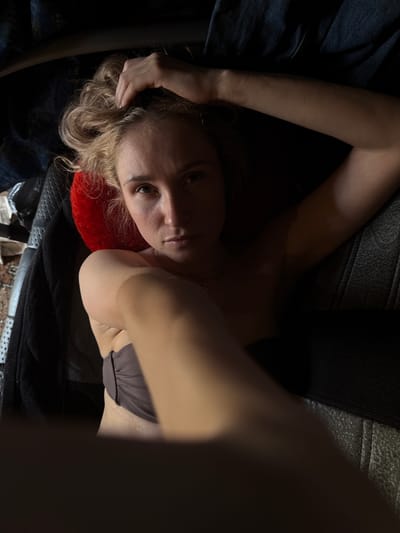 A woman is lying on a dark surface with a red pillow under her head. She is wearing a bra-like top. Her left arm is raised, with her hand near her head. The environment is dark and indistinct. The location is Усть-Тунтор, Russia.