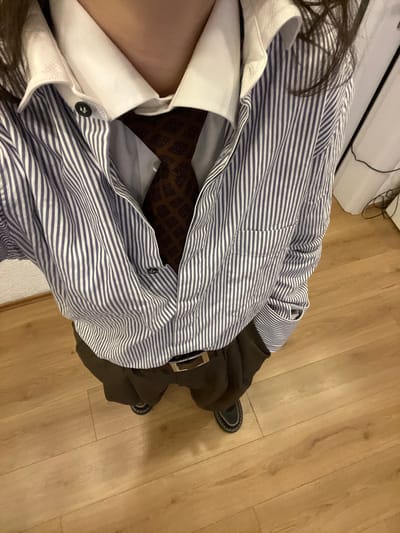 An individual wears several layers of clothing. A white dress shirt with black buttons is visible at the neck. Above that, a blue and white striped shirt is worn. A brown patterned tie peeks from beneath. A brown, belted coat is also part of the ensemble, as well as black shoes. The person stands on a light brown wood floor, adjacent to a white appliance. The camera's angle is overhead, looking down.