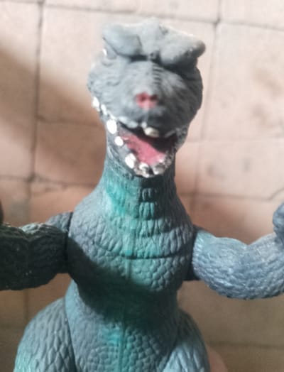 The image shows a close-up, eye-level view of a toy dinosaur figurine. The figurine is predominantly grey with some teal coloration on its chest and neck. It has an open mouth with visible teeth and a pink interior. The background is a blurred brick wall. No specific location in San Lucas Tulcingo, Mexico, is identifiable from the image.