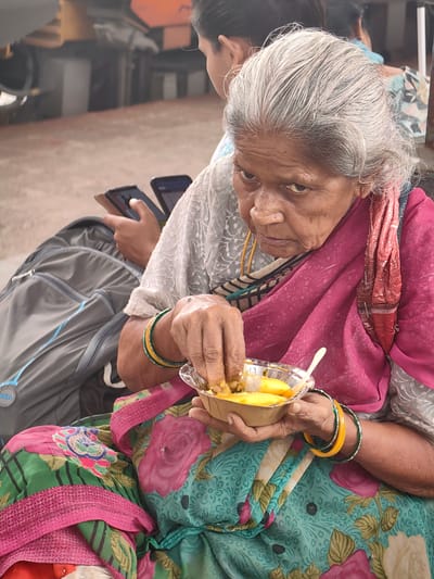 An elderly woman with graying hair, wearing a pink and green floral sari, sits outdoors and eats from a small disposable bowl. Her right hand is in the bowl, picking up food. She is wearing several bangles on her left wrist, including yellow and green ones. A pink scarf is draped around her shoulders, and a grey backpack rests beside her. In the background, another person is partially visible, holding two cell phones. The setting appears to be a public outdoor area, possibly a waiting area or a street scene, given the presence of a backpack and the casual attire of the individuals. The overall scene suggests a moment of everyday life in Itarsi, India.