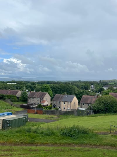 The image presents a wide, slightly elevated view of a residential area situated on a hillside, looking out towards a distant landscape. In the foreground, on the right, is a large expanse of green grass with distinct dirt tracks running through it. At the bottom right, several shipping containers are visible: a blue one with "CARGAS" written on its side, and a green one marked with "P&O", "UAN", "22G1", and "TARE". A small, dark figure, possibly a person, stands near the blue container, and another indistinct dark shape is in the grassy field further back.
The mid-ground features numerous houses, many with cream-colored facades and tiled roofs. Some houses have darker, possibly slate or modern flat roofs, and solar panels are visible on at least one rooftop. These residences are interspersed among abundant green trees, forming a dense canopy that extends into the middle distance. Fences and what appear to be roads or pathways run between the houses. A dark green utility box or shed is located near some properties.
The background shows a hazy distant landscape, suggesting a wide valley or a body of water framed by hills, fading into the atmospheric perspective. The sky is largely overcast, dominated by various shades of grey and white clouds, though hints of clearer blue sky and brighter clouds are visible in certain areas, particularly towards the left. The scene depicts a daytime setting with soft, diffused light, characteristic of a cloudy or partly cloudy day in a temperate climate. The overall impression is a quiet, residential landscape.