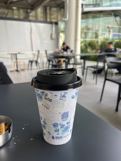 A close-up shot shows a disposable coffee cup on a grey table, likely outdoors. The cup, white with a blue floral design, has a black lid. Text printed on the cup includes "A136 KKM ICE, REGULAR JUWARA BEANS," "fahmi DI," "MAY 27, 14:59," "SCAN HADIAH KEJUTANE," and "HALAL INDONESIA." An ashtray containing cigarette butts is visible in the lower right corner. In the blurred background, chairs and tables suggest an outdoor seating area of a cafe or restaurant. People are visible in the background, suggesting a public space. The location is in the city of Ramunia Dua, Indonesia.
