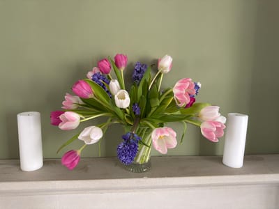 A still-life scene shows a bouquet of tulips and hyacinths in a glass vase, flanked by two white pillar candles. The flowers are in shades of pink, white, and purple, with green stems and leaves. The arrangement sits on a gray shelf, which is presumably a mantelpiece due to its placement against a muted green wall. The setting appears to be indoors, and while the specific location in Malvern Hills, United Kingdom, is not discernible from the image, it is likely a residential interior given the decor. There are no people or actions evident in the photograph, which focuses on the arrangement of the flowers and candles as its subject.
