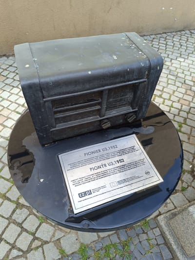 The image depicts a black, weathered metal sculpture of an old radio, specifically a "PIONYR U3,1952," displayed outdoors on a round black pedestal. The pedestal is placed on a cobblestone ground with some grass growing between the stones. A plaque is affixed to the front of the pedestal, providing information about the radio sculpture. The background shows a textured wall and more cobblestone. The sculpture appears to be located in the city of Dzierżoniów, Poland, as indicated by the text on the plaque which mentions "Dzierżoniów" and a project co-financed by the European Regional Development Fund. There are no people visible in the image.