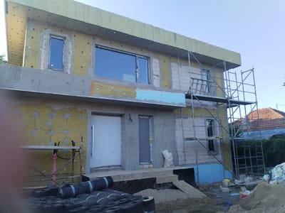 In the city of Dumbrăveni, Romania, a two-story residential building is under construction. The exterior walls are being insulated with yellow mineral wool, which is attached with metal pins. The building has several windows, some of which are already installed, and two doors leading into the structure. A scaffolding system is erected around the building, suggesting ongoing exterior work. Rolls of black roofing material are stacked in the foreground, along with sand and construction debris, indicating that the exterior finishing work, such as roofing and waterproofing, is in progress. The sky is clear and blue, and the surrounding environment appears to be a residential area with trees and other houses visible in the background.