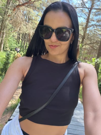 A woman with long dark hair and sunglasses takes a selfie outdoors. She is wearing a black ribbed tank top and has a black strap from a bag across her chest. In the background, there are trees and foliage, suggesting a natural environment. Two people are visible in the distance. The woman appears to be standing on a wooden walkway. The scene depicts a sunny day. The location is the city of Morskoe, Russia.