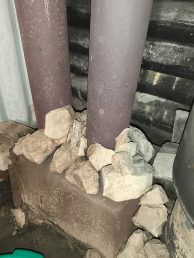 In a dimly lit, possibly outdoor or unheated space in Il'mino, Russia, two large, rusty, maroon-colored pipes stand vertically. They are situated next to a weathered, grey corrugated metal wall. At the base of the pipes, resting on a rough, concrete-like foundation, is a pile of light-colored, irregularly shaped rocks. The ground is a dark, gritty surface. The overall scene suggests a utility area or storage space, possibly exposed to the elements given the presence of rust and natural rock formations.