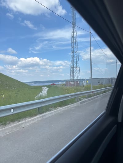 The image depicts a roadside scene with a large metal tower in the background, likely a communication tower. To the left, a grassy embankment slopes upwards, and a metal guardrail runs along the side of the road. In the middle ground, there are several gray modular buildings and a smaller brick structure, all enclosed by a fence. A body of water, possibly a river or a bay, is visible in the distance. A red car is parked near the water's edge, and a person is standing next to it. The sky is blue with scattered white clouds. The scene appears to be in an industrial or semi-rural area. The location is specified as Grebeni, Russia.