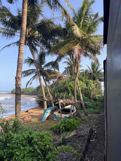 This image depicts a coastal scene in Katukurunda, Sri Lanka, viewed from the side of a moving train. In the foreground, lush green vegetation and palm trees dominate the landscape. A small, overturned boat, painted in blue, white, and green, rests on the sandy shore near a cluster of palm trees and debris. Beyond the shore, the ocean waves gently crash onto the beach under a clear blue sky. In the mid-ground, a sandy beach stretches towards a headland covered in dense green foliage. The right side of the image shows the side of a train carriage, suggesting the perspective of a passenger observing the passing scenery. The overall atmosphere is serene and tropical.