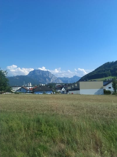 This is a daytime, outdoor, eye-level, medium shot photograph of a rural village in Innergrub, Austria. The foreground is dominated by a field of tall, dry grass, with some patches of greener grass visible. In the middle ground, a cluster of houses and buildings are visible, with a prominent beige and white building on the right side. The background features a large, rugged mountain range under a clear blue sky with scattered white clouds. The overall scene depicts a serene rural landscape with residential structures and natural elements.