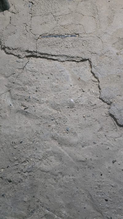 The image displays a close-up, vertical view of a cracked and textured concrete surface. The concrete is light gray with visible aggregate, small stones, and a rough, uneven finish. A prominent, dark horizontal line or embedded object runs across the upper central portion of the image, seemingly integrated within the concrete. The surface exhibits multiple cracks of varying widths and depths, with some areas appearing chipped or eroded. The lighting suggests an outdoor environment, casting subtle shadows that highlight the texture and irregularities of the material. The scene is devoid of any human presence or identifiable landmarks. This concrete surface is located in Yakkabog, Uzbekistan.