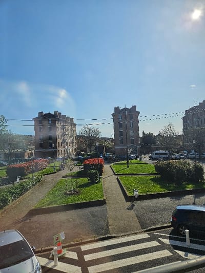 The image depicts an urban outdoor scene in Villeneuve-Saint-Georges, France, under a clear blue sky with direct sunlight, indicated by lens flare in the upper right. The scene is viewed from an elevated perspective, possibly from a window, with reflections or optical artifacts visible, including a prominent horizontal dashed line traversing the mid-ground.
The foreground features a partial view of a silver passenger car on the left and a dark-colored SUV/hatchback on the right. A white pedestrian crosswalk with horizontal stripes spans the width of the road in the immediate foreground. Adjacent to the crosswalk, an orange and white traffic cone with an attached sign and two white bollards are visible.
The mid-ground is dominated by a public square or plaza comprising a network of light-colored concrete pedestrian pathways winding through well-maintained green grassy areas. Several shrubs, some exhibiting reddish foliage, and small trees are interspersed throughout these green spaces. A single individual, a child wearing a light-colored coat, is walking rightward on one of the concrete pathways in the central mid-ground. A white public trash receptacle stands beside a pathway.
In the background, multiple multi-story residential buildings constructed of brick with dark tiled roofs and prominent chimneys are visible. Two particularly tall, slender buildings stand out, one to the left and one to the right of the central axis. Beyond the plaza, paved roads are visible with various parked vehicles, including a white van and several cars. Streetlights are also discernible within the background area.