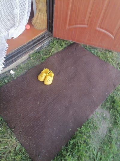A pair of bright yellow children's clogs are placed on a dark brown doormat positioned on the grass outside a wooden door. A white lace curtain is visible to the left of the door, partially obscuring an orange spherical object. A broom leans against the doorway. The scene appears to be in front of a building in the city of Степной, Russia.