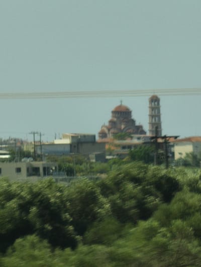 The image captures a view of buildings and a church in Nea Moudania, Greece. In the foreground, there is lush green foliage that is blurred due to motion, suggesting the photo was taken from a moving vehicle. In the middle ground, several buildings are visible, including what appears to be a light-colored structure with windows. To the right of these buildings, a large church with a prominent dome and a tall bell tower is situated. The church is a reddish-brown color and has a distinctive architectural style. Power lines are present in the upper portion of the image, stretching across the sky. The sky itself is a pale blue-gray color. The overall scene depicts a part of the urban landscape of Nea Moudania.