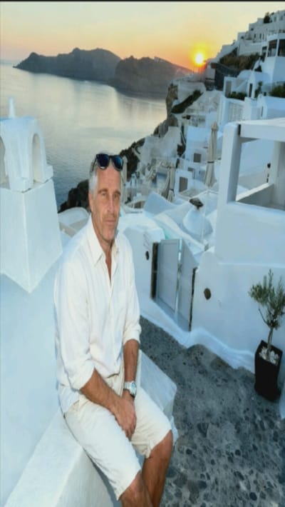 A man, likely in his 50s or 60s, sits on a whitewashed ledge. He is dressed in an unbuttoned white shirt, tan shorts, and a watch on his left wrist. Sunglasses rest on his head. Behind him, the sun sets over a vast body of water and an island. Whitewashed buildings with domed roofs cover the terrain behind the man. A potted plant sits to his right on a dark gray stone walkway. The overall scene conveys a sense of relaxation and wealth.