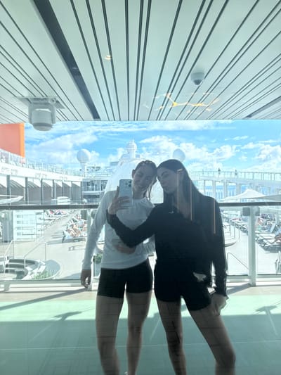 Two females are observed posing for a selfie in front of a large window aboard a cruise ship. The scene is depicted as being in Long Island, Bahamas.
The subject on the left, with light brown hair pulled back, wears a white long-sleeved athletic top and dark athletic shorts. She holds a light-colored smartphone in her right hand, actively taking the photograph, with her left arm around the waist of the other subject. The subject on the right, with long dark hair, wears a black long-sleeved athletic top and dark athletic shorts, with her right arm around the left subject's waist. Both subjects are fair-skinned.
Through the window, the sunny upper deck of a cruise ship is visible. The deck features numerous unoccupied and some occupied lounge chairs, small tables, and white railings. Several indistinct figures are present on the ship's deck and upper levels. The ship's superstructure, including large white communication domes and various architectural elements, is visible against a clear blue sky with scattered white clouds. The setting is outdoors and appears to be in warm weather conditions.
The reflection in the window shows the interior ceiling, which consists of parallel white panels with darker linear accents. A white dome-shaped security camera and a circular white ventilation or smoke detection unit are visible in the upper section of the reflection.