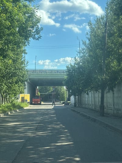 The image depicts a street scene in Gol'yovo, Russia, under a bright, cloudy sky. A wide, paved road dominates the foreground, with strong shadows from trees cast across its surface. On the left, a dense line of lush green trees flanks the road, with glimpses of a sidewalk and a low concrete barrier. Further down the road, a red truck is parked near a yellow truck with some text on its side. To the right of the road, a tall, weathered concrete wall runs parallel to the street, with several trees and lamp posts planted along its edge. An elevated green overpass arches across the scene in the background, supported by concrete pillars. A person is visible walking across the road in the middle distance. The overall impression is a suburban or industrial area on a sunny day.