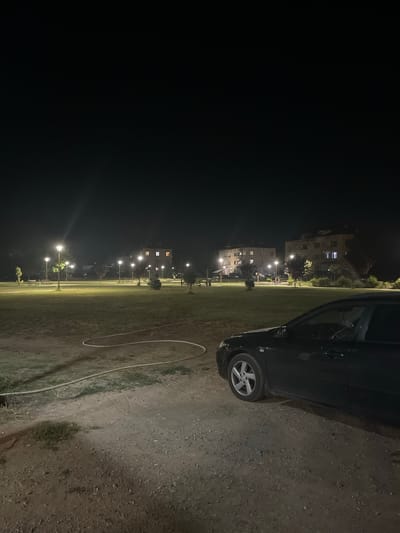 A black sedan is parked on a dirt path in front of a grassy park at night. The park is illuminated by streetlights, with several apartment buildings visible in the background. A white hose is coiled on the ground in the foreground. The image appears to have been taken in Vedrare, Bulgaria.