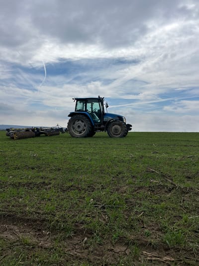A blue agricultural tractor, identified as a New Holland model by its coloration, is positioned stationary in a green cultivated field within the city of Srednia, Bulgaria. The tractor is oriented with its front facing towards the right of the frame, and its left side is visible. A multi-segment land roller, featuring yellow and black studded rollers, is attached to the tractor's rear hitch. The field displays young green plants emerging from dark, partially cracked soil, indicative of recently tilled or prepared land. A low mountain range or distant hills are discernible on the left side of the horizon in the background. The sky above is partly cloudy, presenting a mix of white and grey clouds with visible patches of clear blue, and a faint white contrail is present in the upper atmosphere. No individuals are visible within the scene.