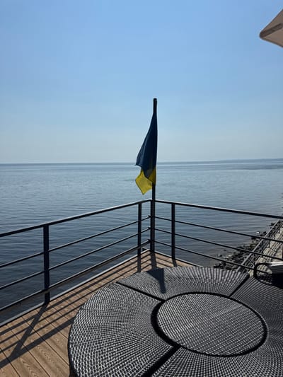 The image presents an outdoor scene overlooking a body of water, identified as the Kyiv Reservoir in Hlibivka, Ukraine. A blue and yellow Ukrainian flag hangs from a dark flagpole, situated near the center-left. The flag appears mostly still or slightly draped. In the foreground, a section of dark, circular woven outdoor furniture, possibly part of a table or seating arrangement, rests on a light brown wooden deck. Dark shadows from the flagpole and a metal railing are cast across the deck, indicating bright sunlight from the upper left. A dark metal railing with multiple horizontal bars runs across the image, separating the deck area from the calm, dark blue water. The far right side of the image reveals a narrow strip of rocky or concrete shoreline at the water's edge. The sky above is clear and bright blue, devoid of clouds. No individuals or animals are visible.
