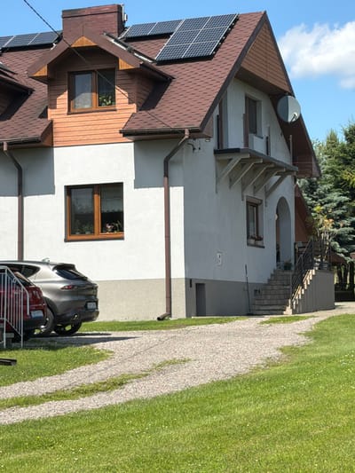 The image shows the exterior of a two-story house with a light gray facade and a brown shingle roof. Solar panels are installed on the roof. There are two cars parked on a gravel driveway in front of the house, one is a dark gray SUV and the other is partially visible in red. A set of concrete stairs leads to the entrance on the right side of the house. The house is surrounded by a green lawn with some small white flowers. In the background, there are tall evergreen trees. The scene appears to be in a residential area, likely in Strzyżowice, Poland, given the context. The sky is blue with scattered white clouds. There are no people visible in the image.