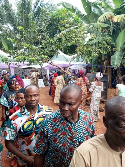 This image captures an outdoor social gathering in Oko Town, Nigeria. In the immediate foreground, three adult males are visible, facing generally forward. The man on the far right is partially seen, wearing a light tan short-sleeved top. To his left, another man wears a short-sleeved shirt with a blue, red, and white geometric patterned design. To his left, a third man wears a white short-sleeved shirt featuring large, multi-colored abstract patterns.
In the midground, a line of approximately ten to fifteen adult individuals, predominantly women, extends towards the left. Most are attired in vibrant, patterned traditional Nigerian dresses and matching headwraps. They appear to be in a queue or procession.
The background reveals a larger gathering area with multiple white canopies or tents, some adorned with purple fabric draping. Numerous individuals are visible under these canopies, with some seated at tables. Lush tropical vegetation frames the scene, featuring large banana plants with broad leaves on the right, palm trees on the left, and various other green trees and shrubs throughout. A prominent tree branch extends horizontally across the midground from the left. The ground surface is visible as reddish-brown earth. A dark, rectangular object resembling a speaker or sound system is positioned upright in the mid-background on the right. The scene is illuminated by natural, diffused light.