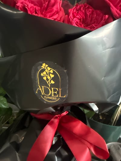 The image shows a bouquet of flowers, likely roses, wrapped in black plastic. A red ribbon is tied around the bouquet. A gold sticker on the wrapper reads "DEL FLOWERS". There is no information to suggest a specific location in Des Plaines, United States.