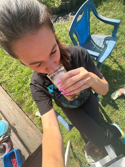 A woman with dark hair, wearing a black t-shirt and black leggings, is sitting outdoors on grass, holding a glass of ice-filled beverage to her lips. Her right hand, with bright pink, pointed nails, is supporting the glass. A blue plastic chair is visible behind her, and to her left, a wooden surface holds containers of red berries, one of which is a blue plastic basket. Her left leg is bent, and a blue shoe with yellow accents is on her left foot. The background consists of green grass and dappled sunlight. The scene appears to be a backyard or park setting. The location is Белая Холуница, Russia.