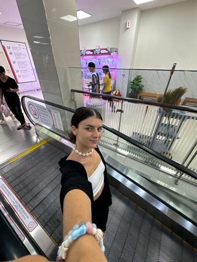 In the city of Phu Thuy, Vietnam, a young woman with dark hair pulled back stands on an escalator, holding her phone to take a selfie. She is wearing a white crop top, a black cardigan, and black pants. A shell necklace adorns her neck, and a beaded bracelet is on her wrist. Behind her, the escalator continues upwards, with other shoppers and an arcade game visible in the background. The escalator is equipped with safety signs and handrails. The overall environment appears to be a modern shopping mall with a clean and well-lit interior.
