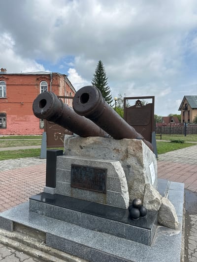 The image depicts a historical monument featuring two large, weathered cannons mounted on a stone pedestal. The cannons are made of dark, rusted metal and are positioned at an upward angle. Below the cannons, the pedestal is made of rough-hewn granite blocks. In front of the granite, a tiered base constructed of polished black and gray granite supports the monument. A bronze plaque is affixed to the granite portion of the pedestal. To the right of the cannonballs, a brown, metal informational plaque with a silhouette of buildings is visible. The monument is situated in an outdoor public space with a paved walkway made of red and grey interlocking bricks. In the background, a two-story red brick building with white-trimmed windows stands to the left, and a smaller, lighter-colored building with a dark roof is visible to the right. A tall, dark green evergreen tree is positioned behind the monument and to the left of the informational plaque. The sky is overcast with patches of blue. The scene is consistent with a memorial site located in the city of Nogornyi, Russia, likely commemorating historical military events or defenses.