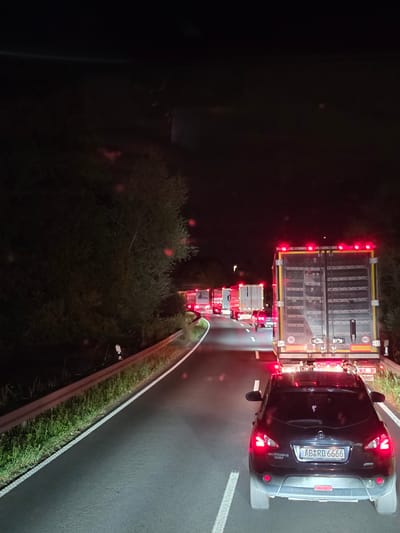 A black Nissan Qashqai is stopped on a highway at night, behind a line of trucks. The car's brake lights are illuminated, and the license plate reads "AB-RD 6666". The highway is dark, with trees and vegetation lining the sides. The road appears wet, suggesting recent rain. The scene is consistent with a traffic jam or accident. This image was taken on the Autobahn A3 near Seligenstadt, Germany.