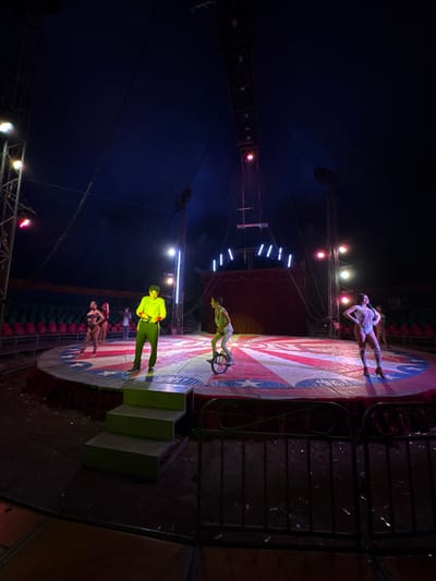 The image captures a circus performance under a tent in Tejalpa, Mexico. The circular stage features a design reminiscent of the American flag. Several performers are present: one balancing on a unicycle, another standing on the stage, and others in various poses. The scene is lit with stage lighting, and a tall structure with rigging is visible, suggesting aerial performances. A set of steps leads up to the stage. Seating is visible around the perimeter. The location appears to be outdoors, within the circus tent.